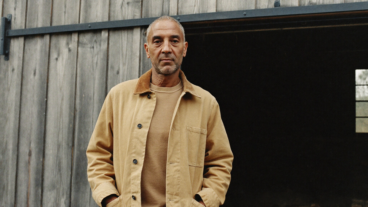 Man in a tan cotton duck chore coat standing in an open barn doorway on an overcast autumn morning