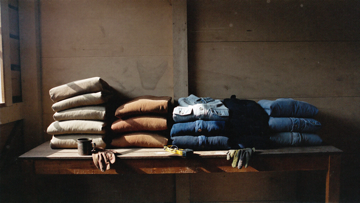 Folded cotton duck and denim jackets stacked on an oak workbench in afternoon light