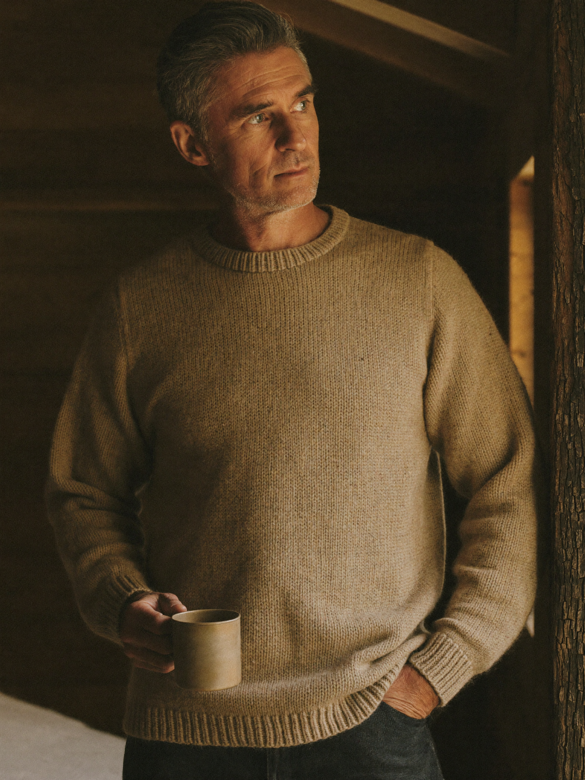 Man in a heavy oatmeal wool crew-neck sweater holding a tin mug in a barn interior